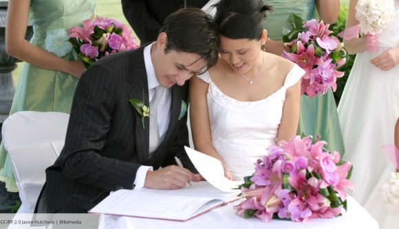 A man in a black suit signs a wedding register beside his new wife, wearing a white dress. Bridesmaids hold a bouquet of pink flowers, and a pink bouquet sits on the table. Image CC-BY-2.0 by Jason Hutchens