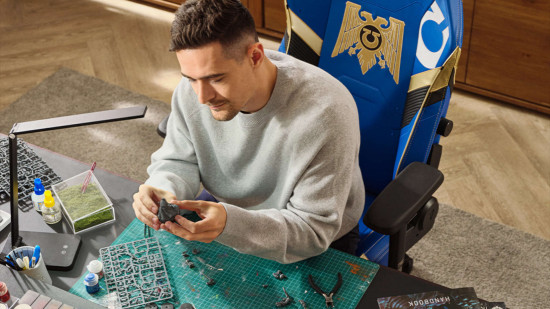 A man sits in a blue Warhammer 40k gaming chair building miniatures at a hobby desk